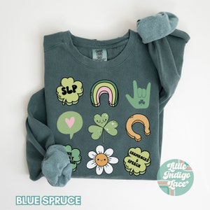 May include: A teal blue sweatshirt with a variety of green shamrock and St. Patrick's Day themed illustrations, including a rainbow, a horseshoe, a heart, a flower, and the text "Shamrocks & Speech". The text "SLP" is also included in a shamrock shape. The sweatshirt is folded in half and the brand name "Little Indigo Lace" is visible in the bottom right corner.