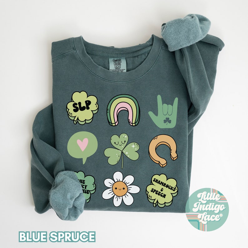 May include: A teal blue sweatshirt with a variety of green shamrock and St. Patrick's Day themed illustrations, including a rainbow, a horseshoe, a heart, a flower, and the text "Shamrocks & Speech". The text "SLP" is also included in a shamrock shape. The sweatshirt is folded in half and the brand name "Little Indigo Lace" is visible in the bottom right corner.