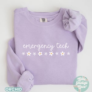 May include: A lavender sweatshirt with the text "emergency tech" in white cursive lettering and a row of white daisies below. The sweatshirt is folded and laid flat on a white surface.