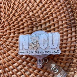 May include: A clear badge reel with the letters "NICU" in white outlined in black, with a teddy bear graphic. The words "neonatal intensive care unit" are written below the letters. The reel has a clear plastic clip.