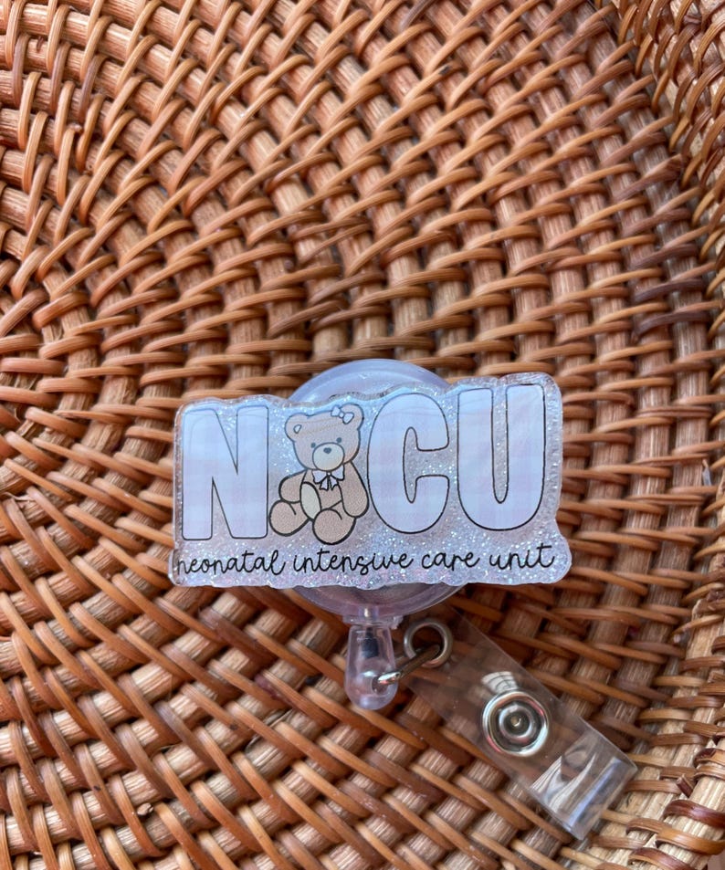 May include: A clear badge reel with the letters "NICU" in white outlined in black, with a teddy bear graphic. The words "neonatal intensive care unit" are written below the letters. The reel has a clear plastic clip.