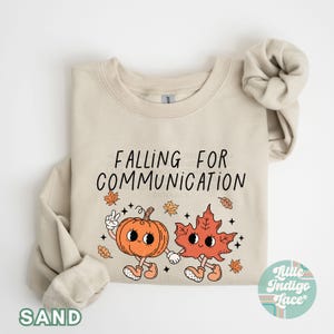 May include: Sand-colored sweatshirt with the text "FALLING FOR COMMUNICATION" and a cartoon pumpkin and leaf holding hands. The pumpkin and leaf have smiling faces and are surrounded by fall leaves. The word "SAND" is printed at the bottom.