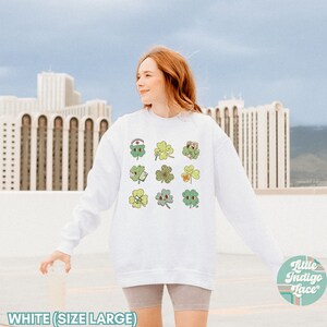 K&ouml;nnte beinhalten: Wei&szlig;er Pullover mit einem Motiv aus neun gr&uuml;nen vierbl&auml;ttrigen Kleebl&auml;ttern, jedes mit einem anderen Design. Die Kleebl&auml;tter sind in einem Raster angeordnet. Der Text "Little Indigo Lace" ist in der rechten unteren Ecke sichtbar.