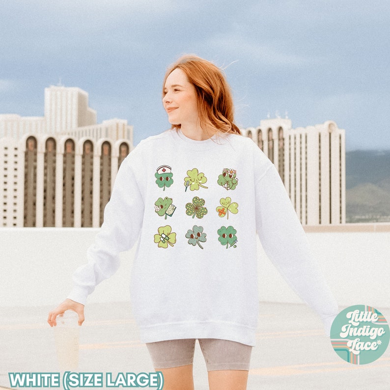 K&ouml;nnte beinhalten: Wei&szlig;er Pullover mit einem Motiv aus neun gr&uuml;nen vierbl&auml;ttrigen Kleebl&auml;ttern, jedes mit einem anderen Design. Die Kleebl&auml;tter sind in einem Raster angeordnet. Der Text "Little Indigo Lace" ist in der rechten unteren Ecke sichtbar.