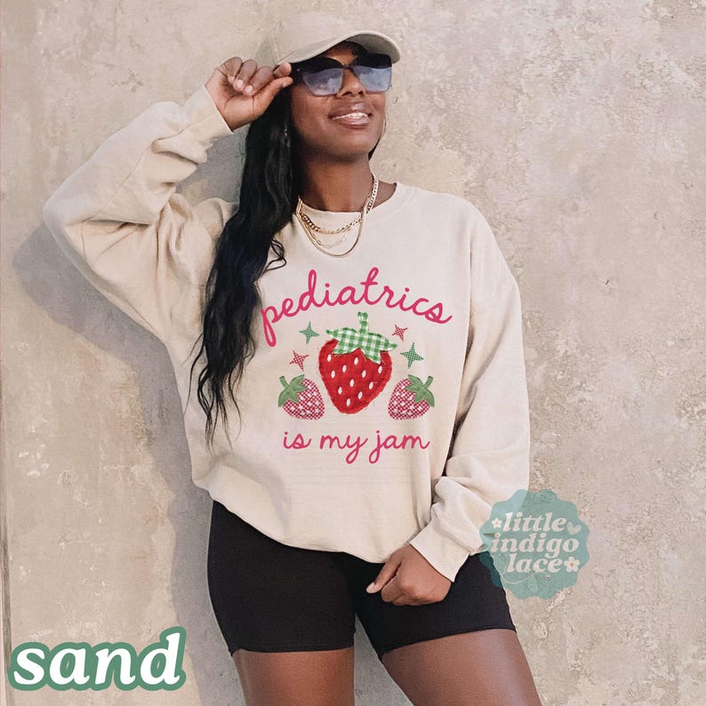 May include: A sand-colored long-sleeve crewneck sweatshirt featuring the red text "pediatrics is my jam" and a graphic of three strawberries with green checkered accents and small star shapes. The sweatshirt is paired with black shorts.