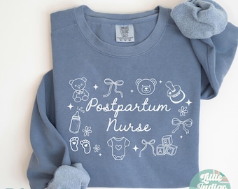 May include: A blue jean colored sweatshirt with the words "Postpartum Nurse" in white script, surrounded by baby-themed illustrations. The sweatshirt has a crew neck and long sleeves. The words "Blue Jean" are at the bottom.