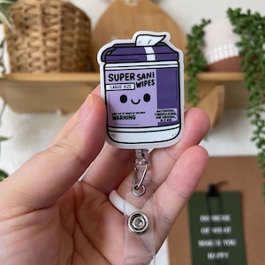 May include: A purple and white badge reel featuring a cartoon illustration of a container of "SUPER SANI WIPES". The text "LARGE SIZE" and "WARNING" are also visible. The reel has a clear plastic strap and a metal clip.