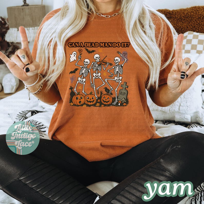 May include: Orange t-shirt with the text "CAN A DEAD MAN DO IT?" and a Halloween-themed graphic. The graphic features dancing skeletons, pumpkins, bats, and a tombstone. The shirt is a comfortable, casual style.