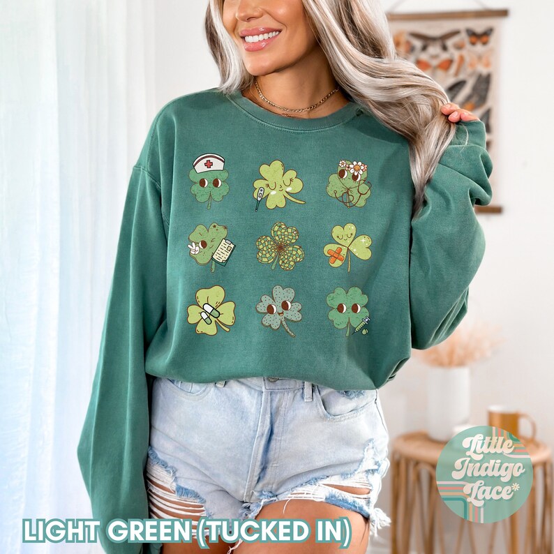 K&ouml;nnte beinhalten: Ein hellgr&uuml;nes Sweatshirt mit einem Aufdruck von neun vierbl&auml;ttrigen Kleebl&auml;ttern, jedes mit einem anderen niedlichen Gesicht und einem kleinen Detail wie einem Verband, einem Thermometer oder einer Pille. Das Sweatshirt ist in die Hose gesteckt und das Model tr&auml;gt Jeans-Shorts.