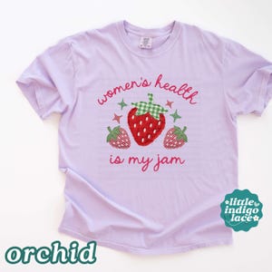 May include: A light purple t-shirt featuring a graphic of three red strawberries with green gingham leaves. The text "women's health is my jam" is printed in pink cursive script above and below the strawberries, accented by small star shapes.
