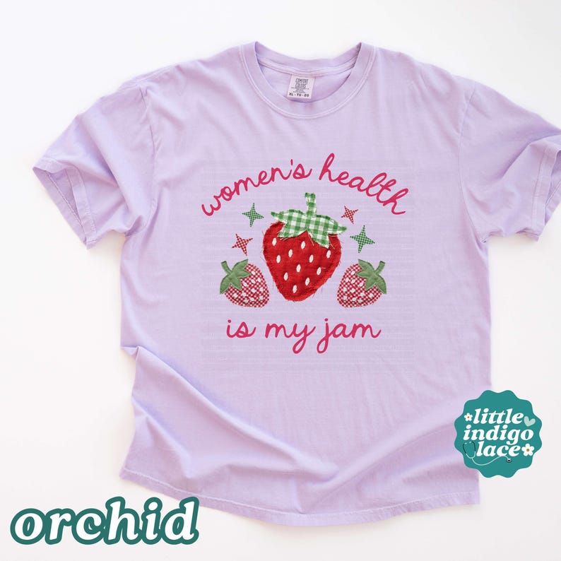 May include: A light purple t-shirt featuring a graphic of three red strawberries with green gingham leaves. The text "women's health is my jam" is printed in pink cursive script above and below the strawberries, accented by small star shapes.