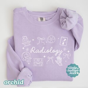 May include: Orchid-colored sweatshirt with white Radiology graphics, including medical equipment and text. The sweatshirt has a crew neck and long sleeves. The brand name "Comfort Colors" is visible on the tag.