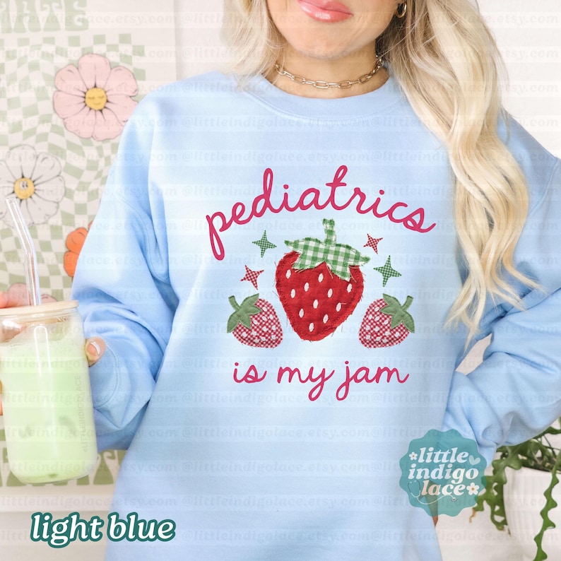 May include: A light blue crewneck sweatshirt featuring the red text "pediatrics is my jam" and a graphic of three strawberries with green checkered leaves. The central strawberry is larger than the two smaller strawberries on either side.