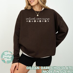 May include: A dark chocolate brown sweatshirt with the text "critical care nurse" and a row of white flower graphics. The model is wearing a size XL.