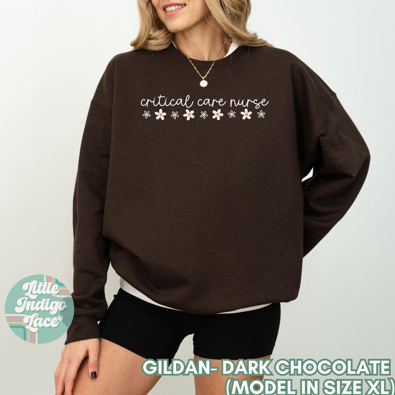 May include: A dark chocolate brown sweatshirt with the text "critical care nurse" and a row of white flower graphics. The model is wearing a size XL.