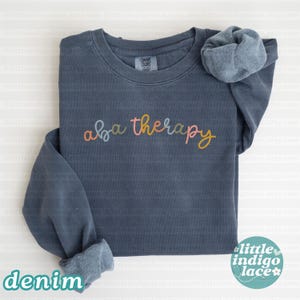K&ouml;nnte beinhalten: Ein denimblaues Comfort Colors Sweatshirt mit dem Schriftzug "aba therapy" in farbenfroher Schrift. Das Sweatshirt hat eine lockere Passform und gerollte B&uuml;ndchen. Das Wort "denim" ist unten aufgedruckt.