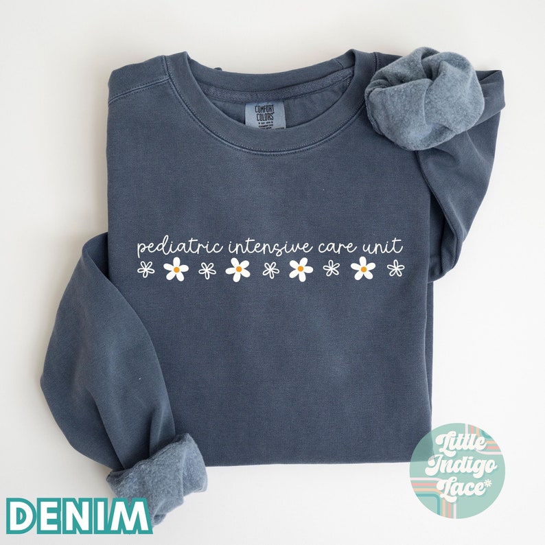 K&ouml;nnte beinhalten: Ein dunkelblauer Pullover mit dem Text "pediatric intensive care unit" und einer Reihe wei&szlig;er G&auml;nsebl&uuml;mchen mit gelben Zentren. Der Pullover ist in der H&auml;lfte gefaltet und der Markenname "Little Indigo Lace" ist in der rechten unteren Ecke sichtbar.