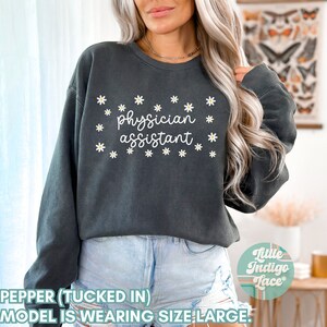 May include: A dark gray sweatshirt with a white daisy print and the text "physician assistant" in white. The model is wearing the sweatshirt tucked in with blue denim shorts.