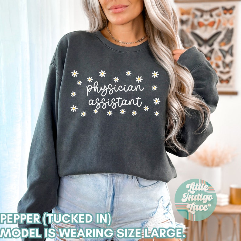 May include: A dark gray sweatshirt with a white daisy print and the text "physician assistant" in white. The model is wearing the sweatshirt tucked in with blue denim shorts.