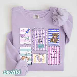 May include: A lilac-colored sweatshirt with a crew neck and long sleeves. The front features several framed illustrations of baby-related items, including a pacifier, diaper, baby bottle, and a teddy bear. Text on the shirt reads "labor and delivery surgical tech" and "it's a good day to catch babies."