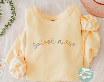 May include: A yellow sweatshirt with the text "school nurse" in colorful lettering. The sweatshirt is folded and laid flat on a white surface.