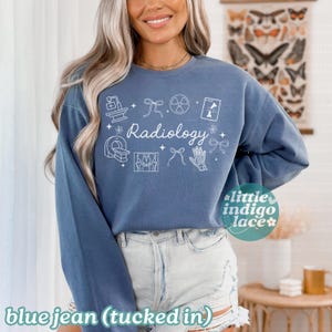May include: A blue jean colored long-sleeve sweatshirt with the word "Radiology" in white script. The shirt features white line drawings of medical imaging equipment and symbols. The text "little indigo lace" is in the lower right corner.