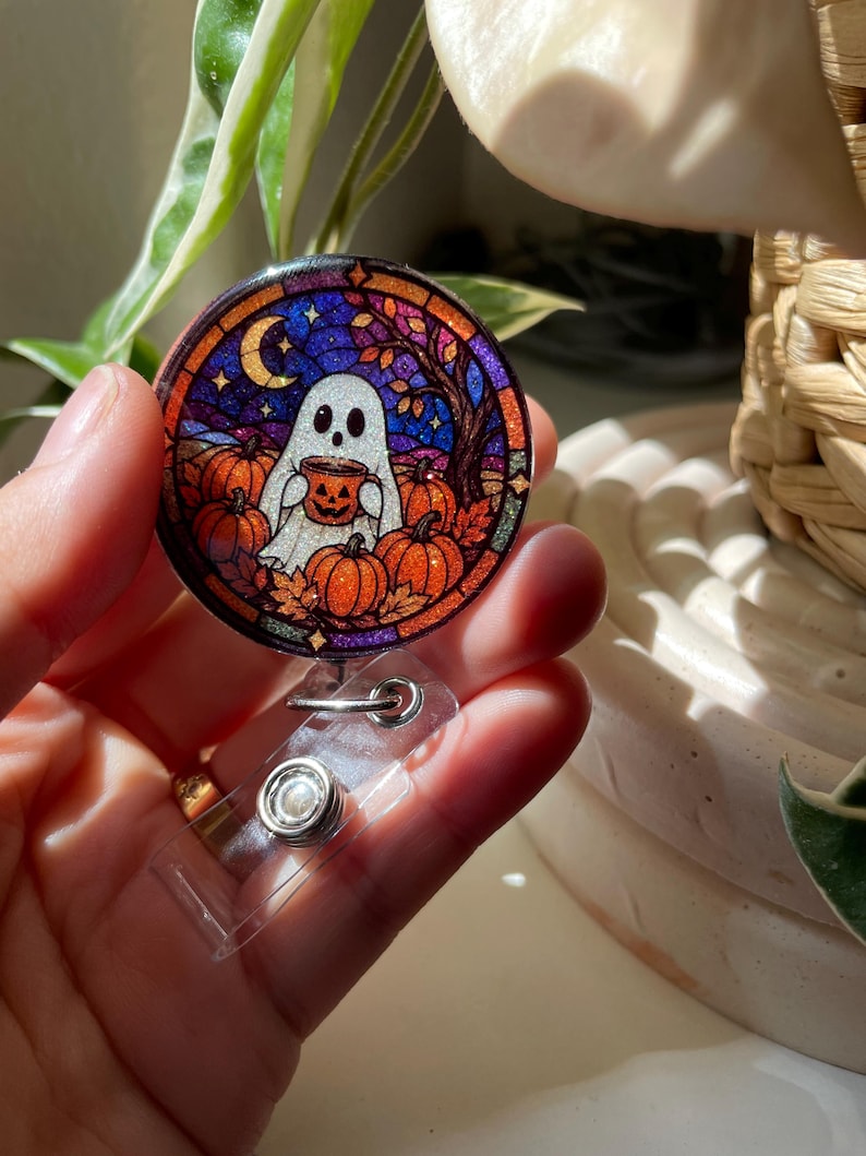 May include: A circular badge reel featuring a stained-glass style illustration of a white ghost holding a jack-o'-lantern mug. The scene includes orange pumpkins, autumn leaves, a crescent moon, and a dark tree against a deep blue night sky.
