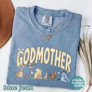 May include: A blue short-sleeve t-shirt with a crew neckline and a Comfort Colors label. The front features the yellow text "GODMOTHER" above a graphic of various cartoon animals including a bear, a tiger, and a donkey, with a gold necklace.