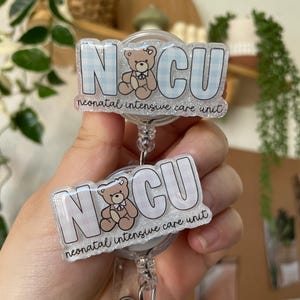 May include: A clear badge reel featuring the letters "NCU" in a light blue gingham pattern, a teddy bear illustration, and the words "neonatal intensive care unit". The reel is held by a clear plastic clip.