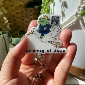 May include: A white acrylic badge reel featuring a green frog wearing a gold crown and blue scrubs, holding a skeletal X-ray while riding a white stork. The text "we x-ray at dawn" is printed in black at the bottom of the badge reel design.