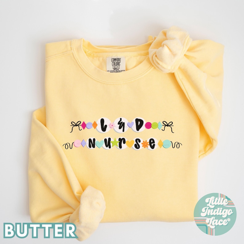 K&ouml;nnte beinhalten: Ein gelber Pullover mit den Worten "L & D NURSE", die in bunten Buchstaben und Formen ausgeschrieben sind. Der Pullover ist gefaltet und hat ein gelbes Haargummi am &Auml;rmel.