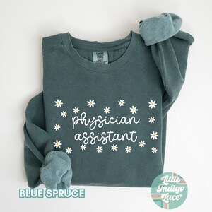 May include: A teal blue sweatshirt with the text "physician assistant" in white script surrounded by white daisies. The sweatshirt is folded in half and the brand label is visible.