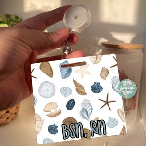 May include: A square badge reel featuring a white background with a repeating pattern of various seashells and starfish in shades of blue, tan, and white. The text BSN. RN is printed in a bold, light blue font across the bottom of the badge.