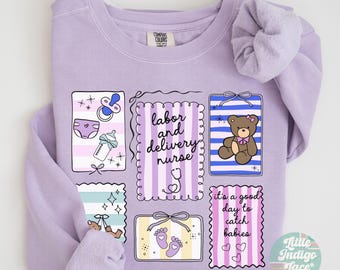 Op de afbeelding: Orchidee-kleurige sweatshirt met een ontwerp met baby-gerelateerde illustraties en tekst. Het ontwerp bevat een fopspeen, luier, babyfles, teddybeer, voetafdrukken en de zinnen "labor and delivery nurse" en "it's a good day to catch babies."