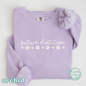 May include: Orchid-colored sweatshirt with the text "future dietitian" in white script, decorated with small daisy graphics. The sweatshirt is a Comfort Colors brand and features a crew neck and long sleeves.
