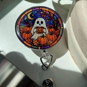 May include: A circular badge reel featuring a glittery stained-glass style illustration of a white ghost holding a jack-o'-lantern mug, surrounded by orange pumpkins, autumn leaves, a crescent moon, and a dark blue night sky background.