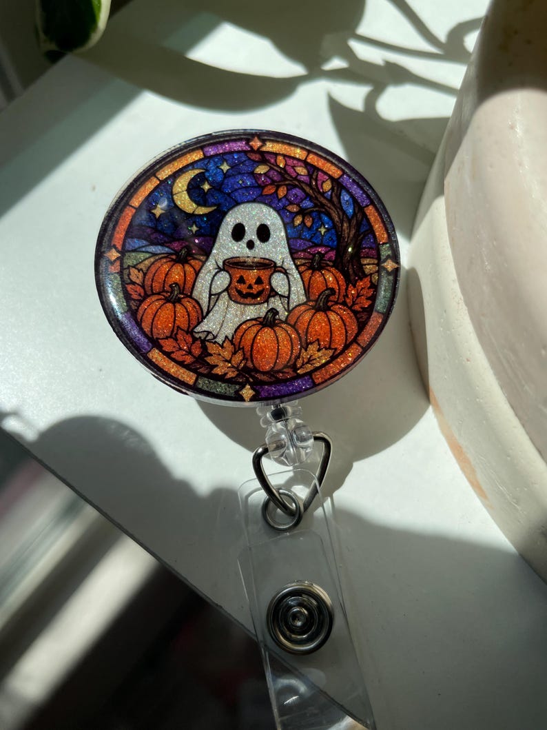 May include: A circular badge reel featuring a glittery stained-glass style illustration of a white ghost holding a jack-o'-lantern mug, surrounded by orange pumpkins, autumn leaves, a crescent moon, and a dark blue night sky background.