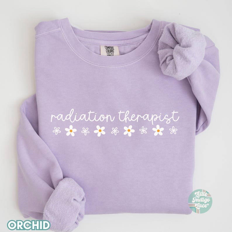 May include: A lavender sweatshirt with the text "radiation therapist" and a row of white daisies printed on the front.