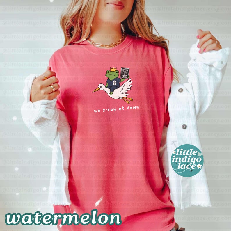 May include: A coral-colored short-sleeve t-shirt featuring a graphic of a green frog wearing a gold crown and a blue medical scrub top, riding a white stork while holding an x-ray. The text below the graphic reads, we x-ray at dawn.