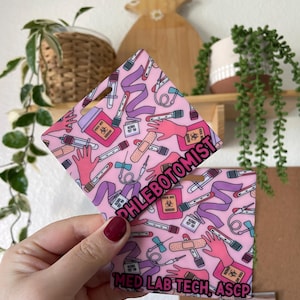 Peut inclure: Badge rose et violet à motifs avec le texte "Phlebotomist" et "Med Lab Tech, ASCP". Le badge est recouvert d'images médicales, notamment des tubes à essai, des bandages et une main gantée.