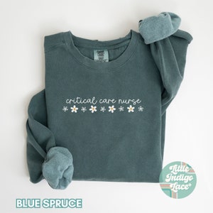May include: A teal blue sweatshirt with the text "critical care nurse" and a row of white daisies. The sweatshirt is folded in half and the brand tag is visible.