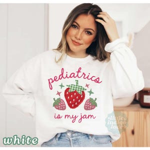 May include: A white long-sleeve sweatshirt featuring the pink text "pediatrics is my jam" and a central graphic of three strawberries. The strawberries are designed with red and green checkered patterns and small green star accents.