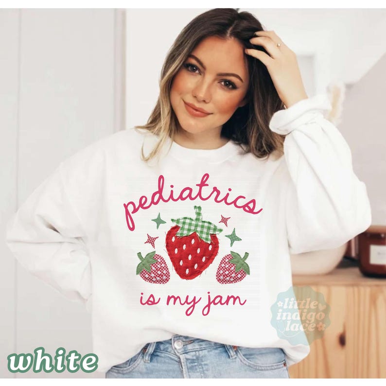 May include: A white long-sleeve sweatshirt featuring the pink text "pediatrics is my jam" and a central graphic of three strawberries. The strawberries are designed with red and green checkered patterns and small green star accents.