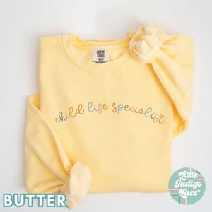 May include: A yellow sweatshirt with the text "child life specialist" in rainbow colors. The sweatshirt is folded and has a scrunchie on top.