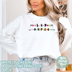 K&ouml;nnte beinhalten: Wei&szlig;er Langarm-Pullover mit dem Text "Love & Do Nurse" in bunten Buchstaben und einem Herz-Design. Das Model tr&auml;gt den Pullover in eine blaue Jeans gesteckt.