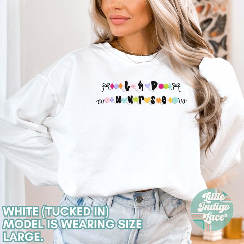 K&ouml;nnte beinhalten: Wei&szlig;er Langarm-Pullover mit dem Text "Love & Do Nurse" in bunten Buchstaben und einem Herz-Design. Das Model tr&auml;gt den Pullover in eine blaue Jeans gesteckt.