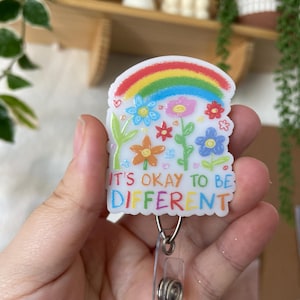 Peut inclure: Un porte-badge en plastique blanc avec un arc-en-ciel, des fleurs et le texte "It's okay to be different". Le porte-badge est doté d'un clip rétractable.