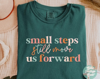 May include: A teal blue t-shirt with the text "small steps still move us forward" in pink, orange, and peach colors. The text is in a handwritten font.