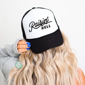 May include: A black and white trucker cap featuring a foam front panel with the text Raisin' HELL printed in black script and block lettering. The cap includes a black peak and a mesh back panel, designed with a structured crown and an adjustable snapback.