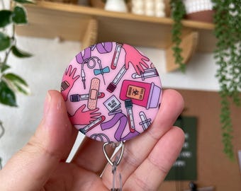 Puede incluir: Un portagafete redondo de color rosa con un patrón de suministros médicos, que incluyen tubos de ensayo, vendas y una jeringa. El portagafete está unido a un carrete retráctil de gafete plateado.